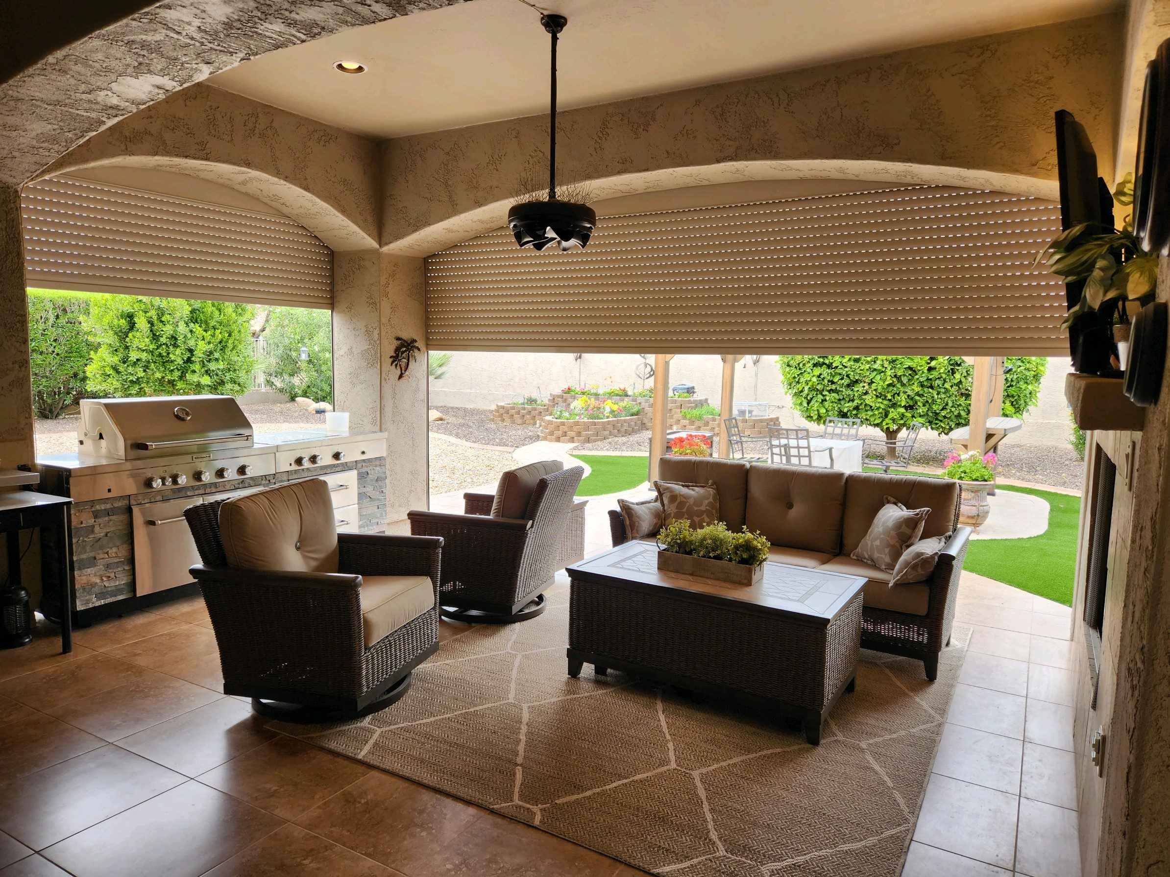 A covered outdoor living space featuring two wicker chairs and a coffee table with a plant. In the background, a barbecue grill and a seating area are visible, with greenery and landscape design visible through the open windows.
