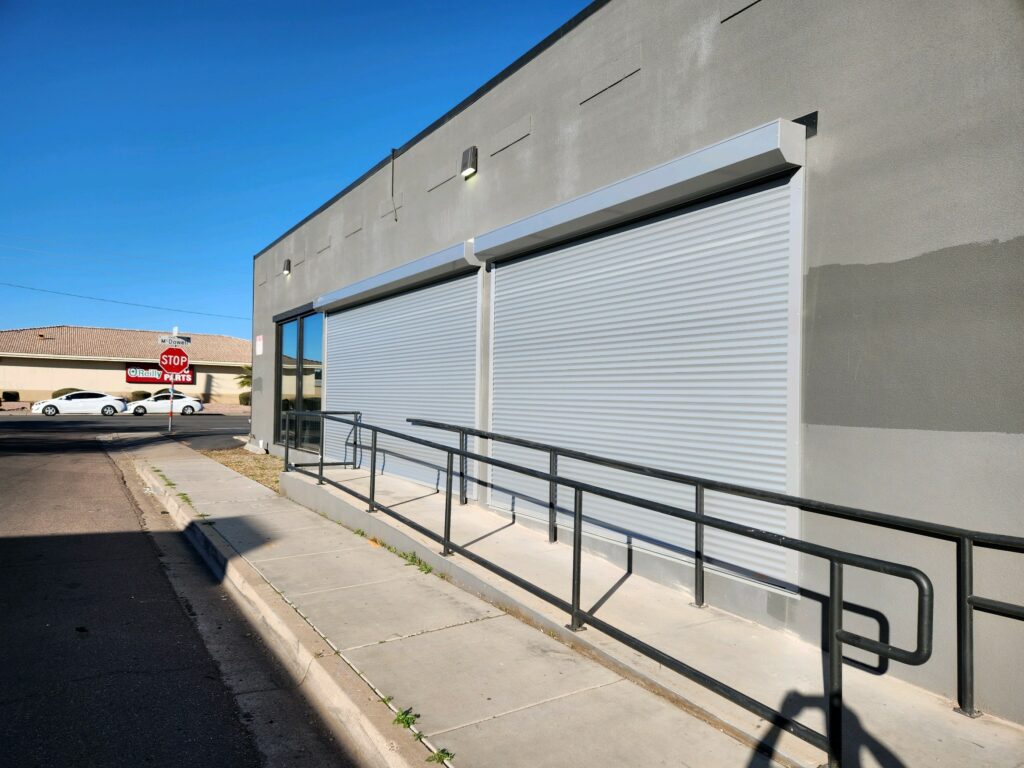 Exterior view of a commercial building with two closed roll-up doors and a wheelchair-accessible ramp. A stop sign is visible in the background, along with parked cars.