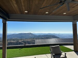 A view from a covered patio showing a clear blue sky and rolling mountains in the background. A paved area with artificial grass and a reclining chair is visible in the foreground.