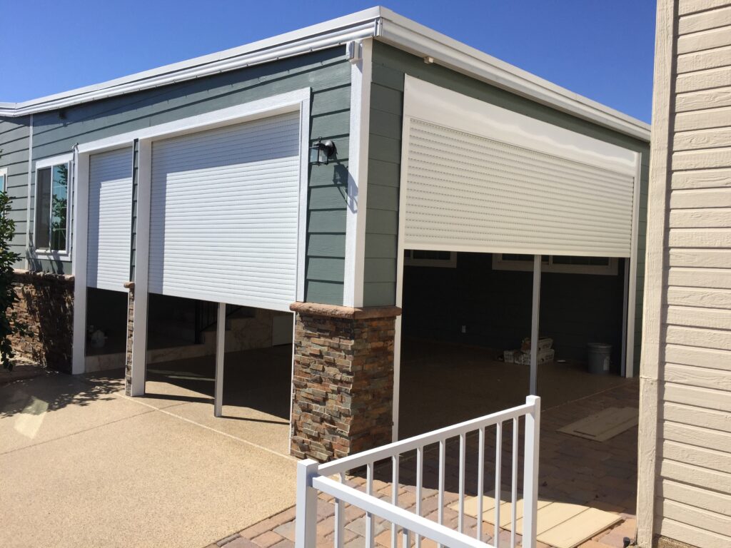 A residential garage with closed rolling shutters on the front, set against a light green exterior wall. The entrance is framed by stone accents, and a concrete pathway leads up to a white metal railing in the foreground.