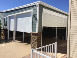A residential garage with closed rolling shutters on the front, set against a light green exterior wall. The entrance is framed by stone accents, and a concrete pathway leads up to a white metal railing in the foreground.