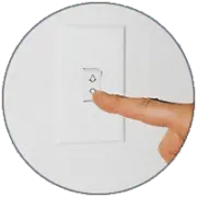 A close-up image of a hand pressing a white light switch on a wall. The switch has a small indicator light and is surrounded by a circular frame.