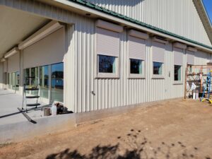 Exterior view of a building with metal siding, featuring several windows and roll-up shutters. A partially finished patio area is visible, with construction tools and materials nearby.