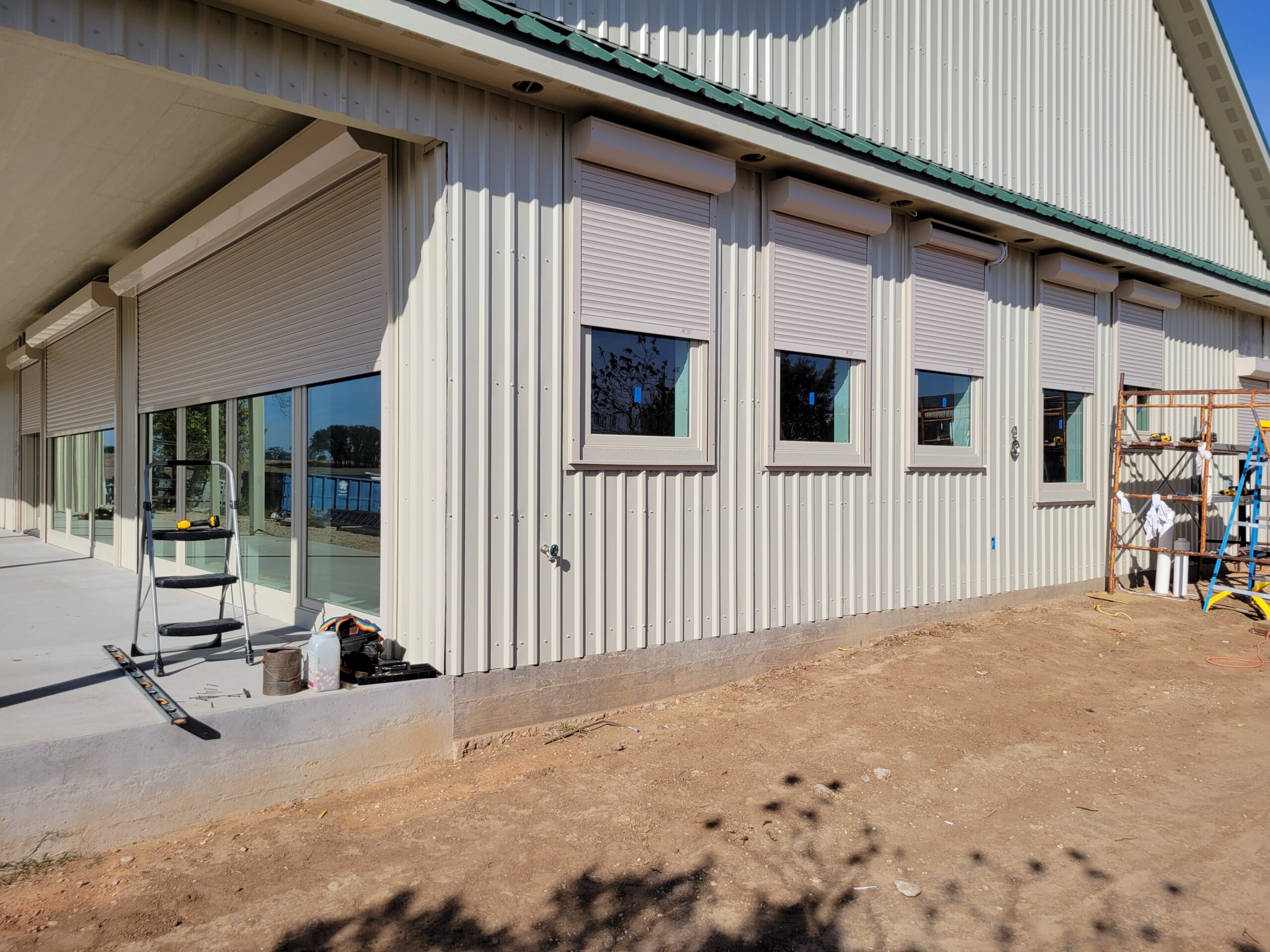 Exterior view of a building with metal siding, featuring several windows and roll-up shutters. A partially finished patio area is visible, with construction tools and materials nearby.