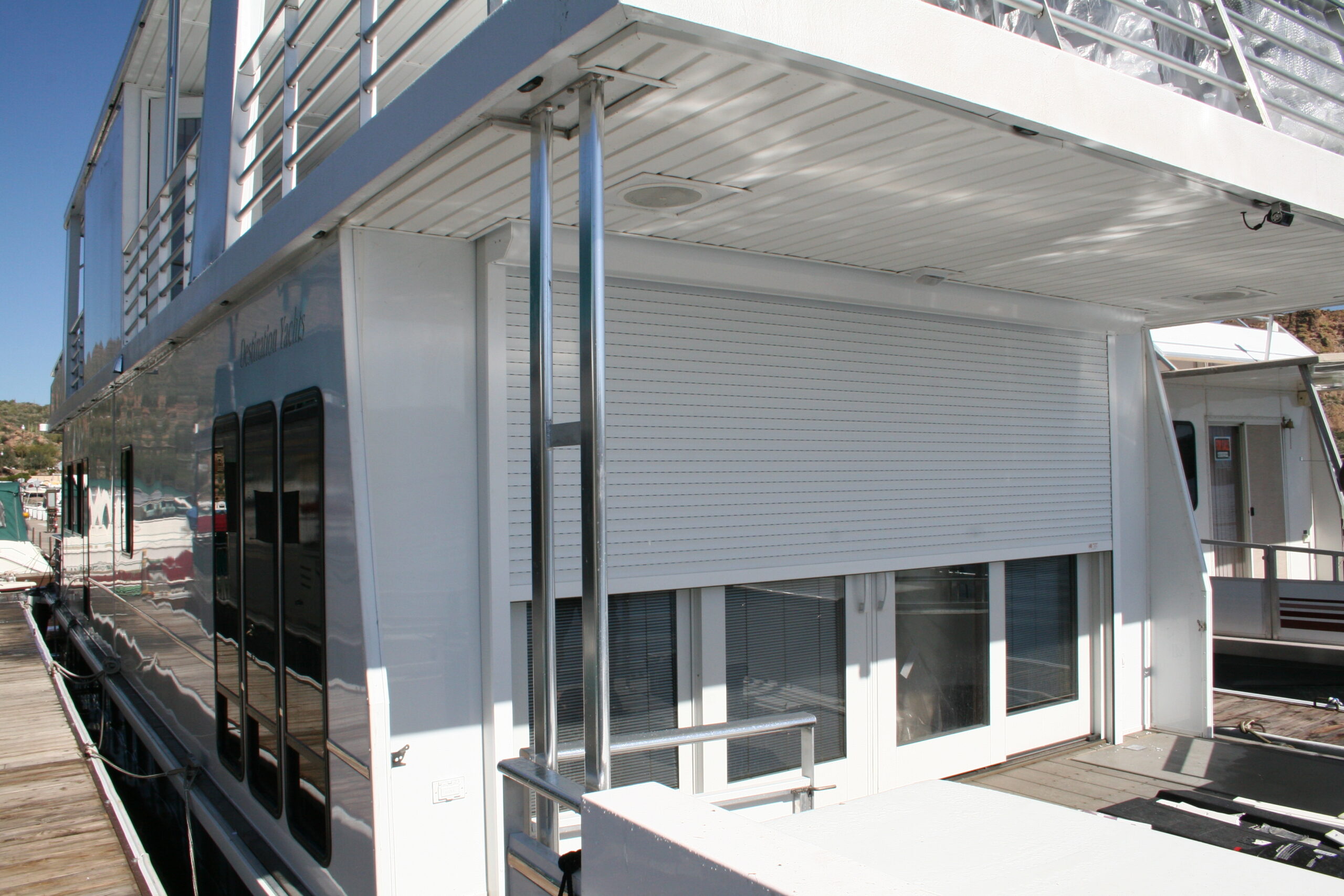 A modern houseboat featuring a large, closed roller shutter covering the entrance. The structure has large windows along the side and is docked at a marina with clear blue skies above.
