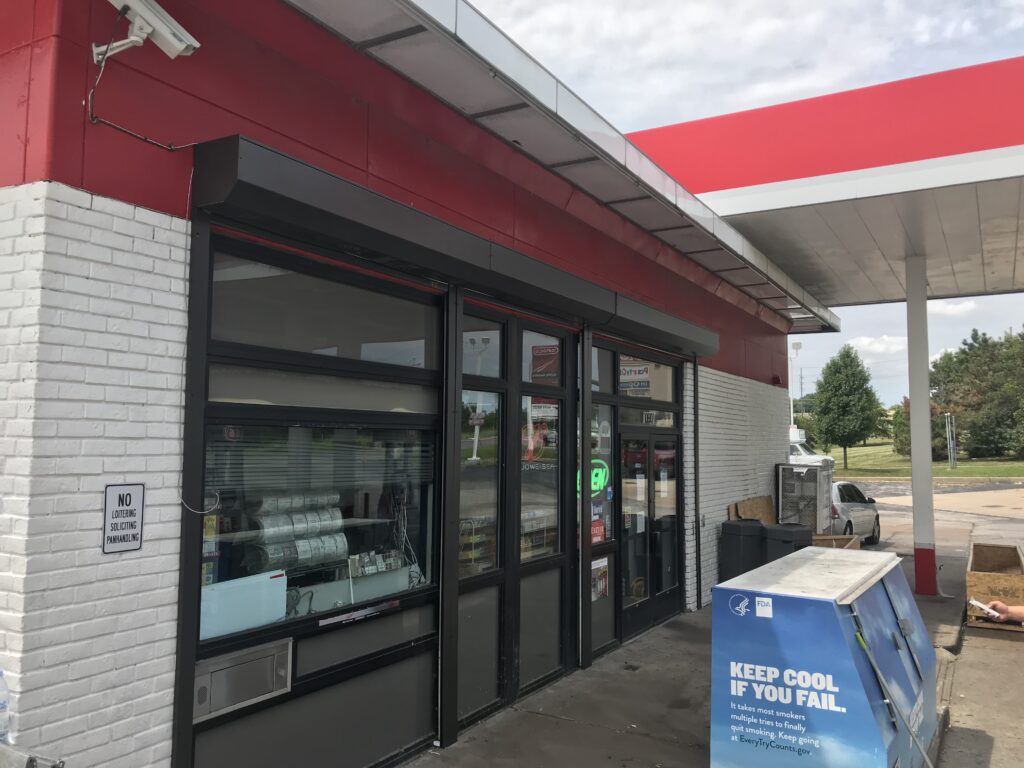 Exterior view of a convenience store with large glass windows and a prominent red roof. A sign indicating 'No Alcohol' is visible next to the entrance, and a blue recycling bin is positioned nearby.