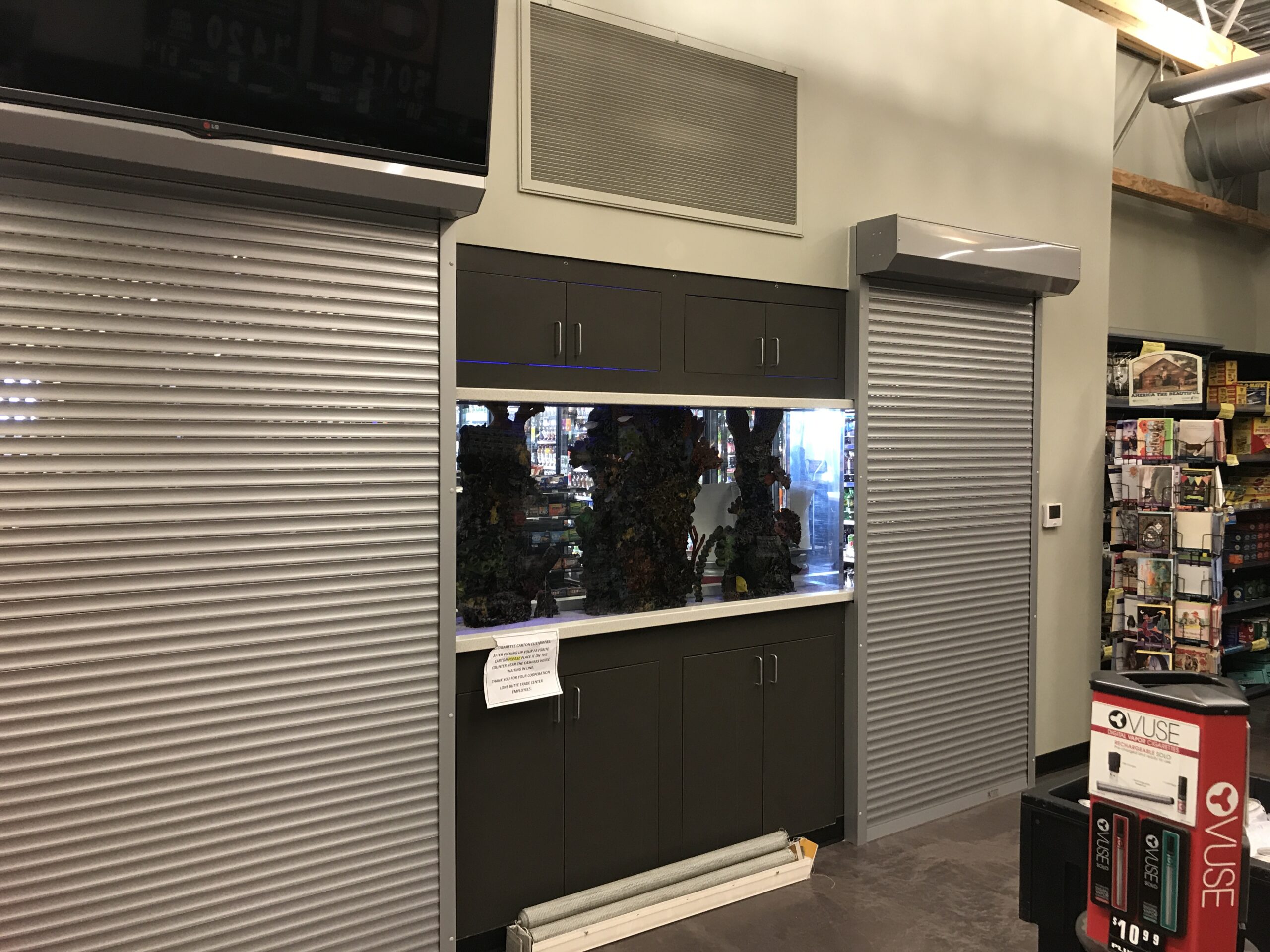 Interior view of a store featuring a gray panel wall with storage cabinets below and a shiny metal surface above. A window with reflection is visible in the center, flanked by metallic rolling doors. A variety of products can be seen on a nearby shelf.
