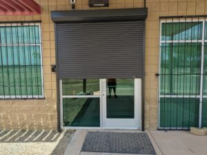 A building entrance featuring a rolling security shutter partially closed over a glass door. The door is framed by metal grilles on either side, with a textured entrance mat in front.