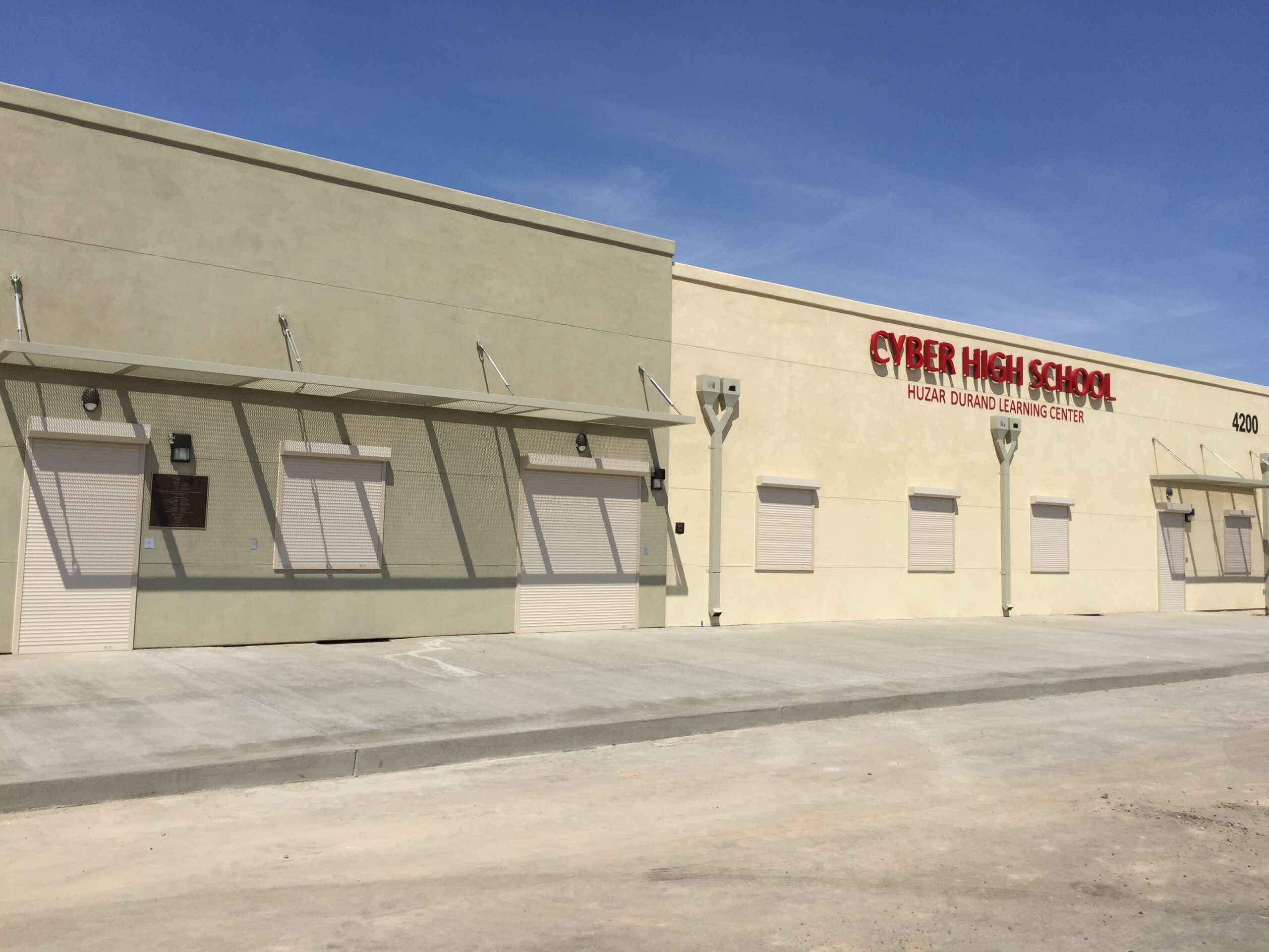The exterior of Cypher High School, featuring a light-colored building with closed shutters. The school's name is displayed in bold red letters above the entrance, under a clear blue sky.