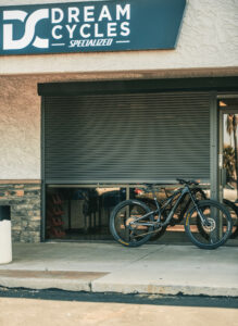 A black mountain bike is parked in front of a shop with a sign that reads 'Dream Cycles Specialized.' The store has a closed shutter and a stone wall design.
