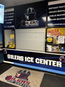 A service counter at Oilers Ice Center featuring a closed shutter, promotional signs for birthday parties and skating events, and a logo with a hockey player. A welcome mat is visible in front of the counter.