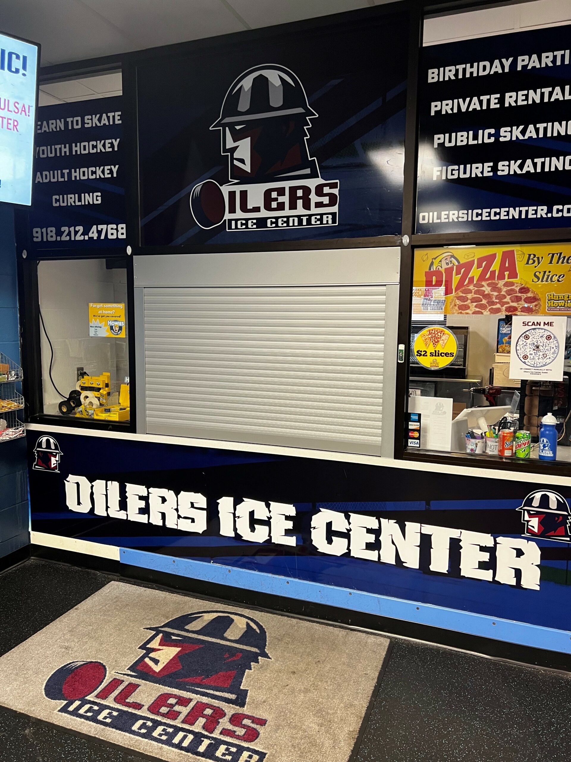 A service counter at Oilers Ice Center featuring a closed shutter, promotional signs for birthday parties and skating events, and a logo with a hockey player. A welcome mat is visible in front of the counter.