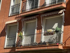 Four windows on a brick building with closed roller shutters and decorative balconies. Each balcony has potted plants, including green foliage and flowers, under white striped awnings.