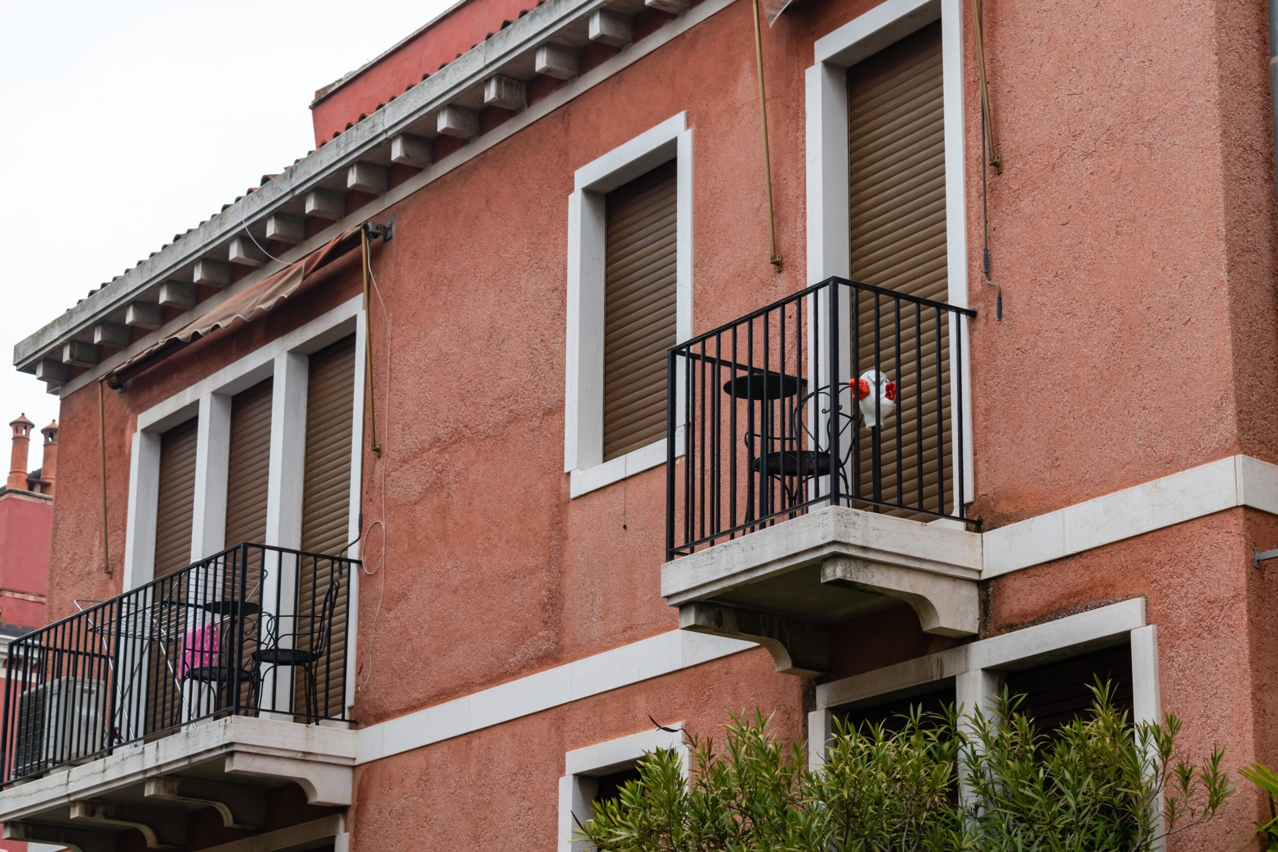 A pink-stucco building with closed window shutters and two balconies. One balcony has a small table and two chairs, while the other displays a pink item. Greenery is visible below.