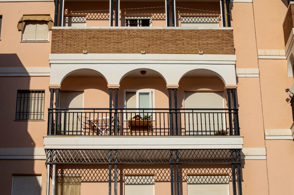 Almunecar,,Spain,-,October,12,,2023:,Walking,On,The,City A peach-colored building featuring two balconies with arched railings. One balcony has a potted plant and a chair visible. The windows above are covered with blinds, and the overall architecture has a modern style.