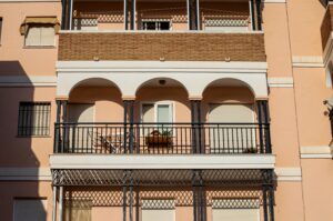 A peach-colored building featuring two balconies with arched railings. One balcony has a potted plant and a chair visible. The windows above are covered with blinds, and the overall architecture has a modern style.