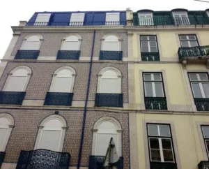 A building facade featuring a combination of blue and beige tiles, with several windows and ornate balconies. The ground level has a white umbrella visible in front, and the structure shows a mix of architectural styles.