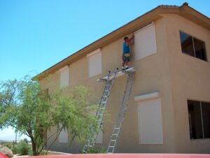 A person is on a ladder, installing window shutters on a two-story beige building. The setting features clear blue skies and a green tree in front.