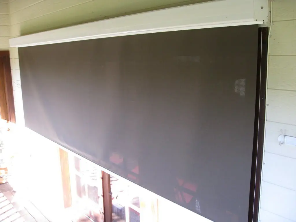 A dark, retractable screen shade is mounted above a window in a wooden frame, partially blocking sunlight from entering a room.
