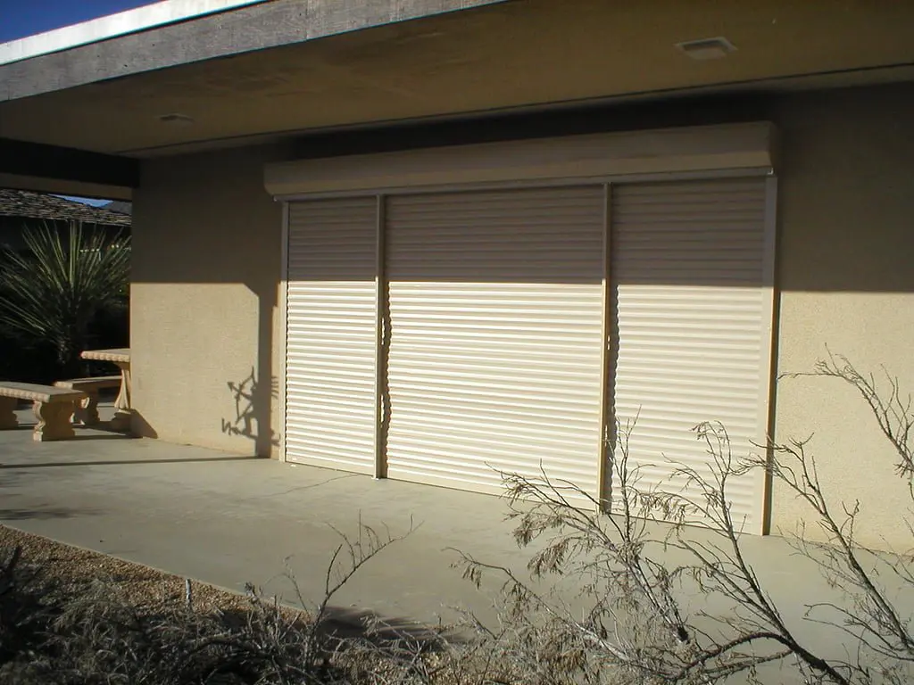 A building exterior featuring two roll-up security shutters closed on a concrete surface, with sparse vegetation and a picnic table in the foreground.