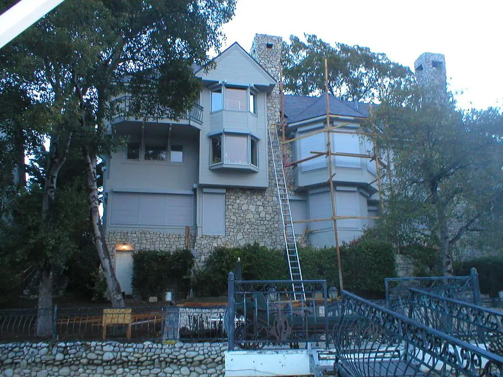 A three-story house with a stone facade and gray siding, featuring multiple windows. A ladder leans against the side, and scaffolding is visible on one section. Surrounding trees provide shade.