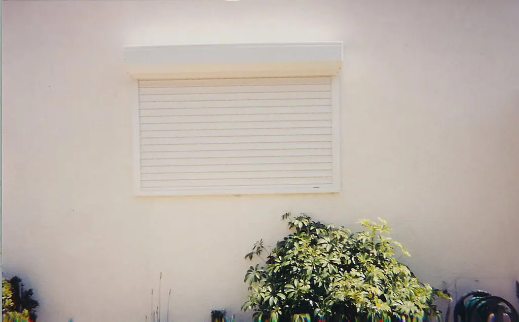 A closed window with horizontal slats, set into a cream-colored wall, surrounded by green plants and a garden hose.