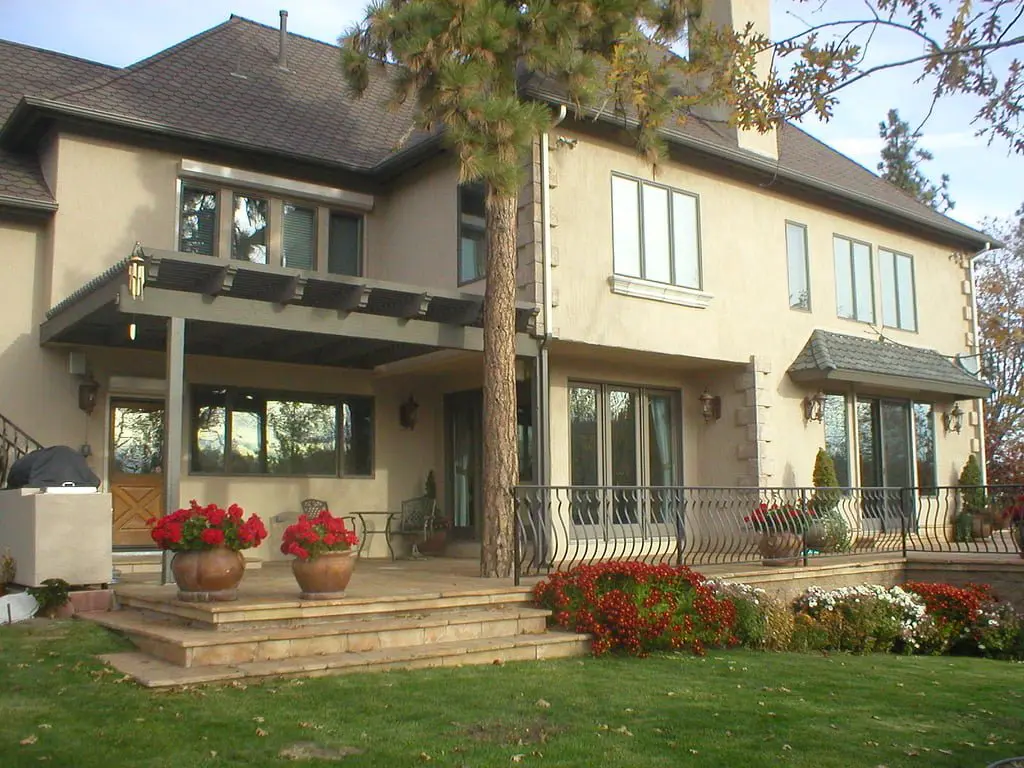 A two-story beige house with large windows, a covered porch, and a balcony. Flower pots with red flowers are placed on the steps, and a green lawn surrounds the property.