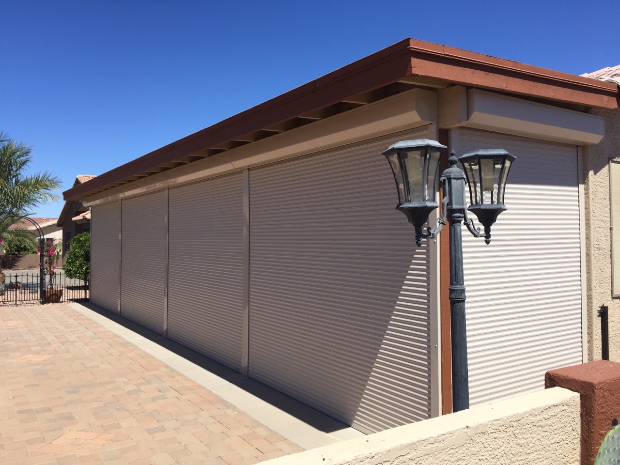 A decorative residential wall with three closed metal shutters and two lamp posts on the side, set against a clear blue sky. The surrounding area features paved flooring and a palm tree in the background.
