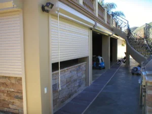 Exterior view of a property featuring two closed roll-up garage doors beside a stone wall, with a pathway leading to a staircase and a small toy car visible nearby.