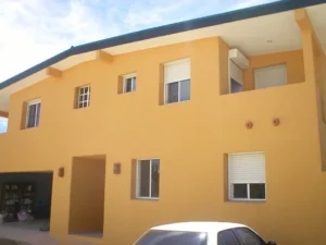 A two-story orange house with multiple windows and roll-down shutters. The garage entrance is visible, along with a car parked in front. The architecture features a sloped roof and a clean, modern design.