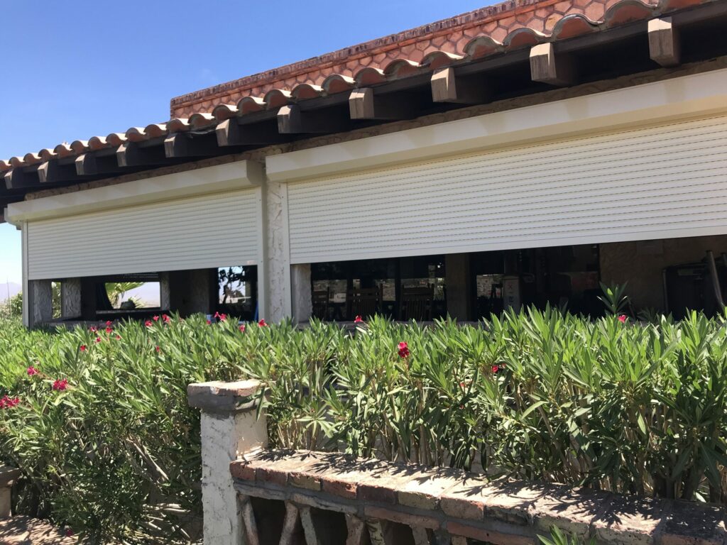 A house with a tiled roof features closed roller shutters on large windows, surrounded by green foliage and blooming pink flowers. The area is sunny and clear.