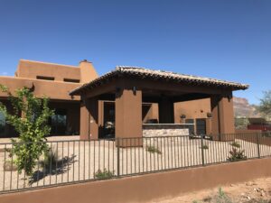 A brown stucco house with a tiled roof and stone bar area on a patio, surrounded by a metal fence. The background features clear blue skies and desert landscaping.