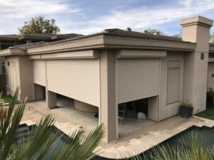 A modern house exterior featuring beige walls and a flat roof. The garage is fitted with a large rolling shutter door. In the foreground, there is a landscaped area with plants and a swimming pool, partially visible.