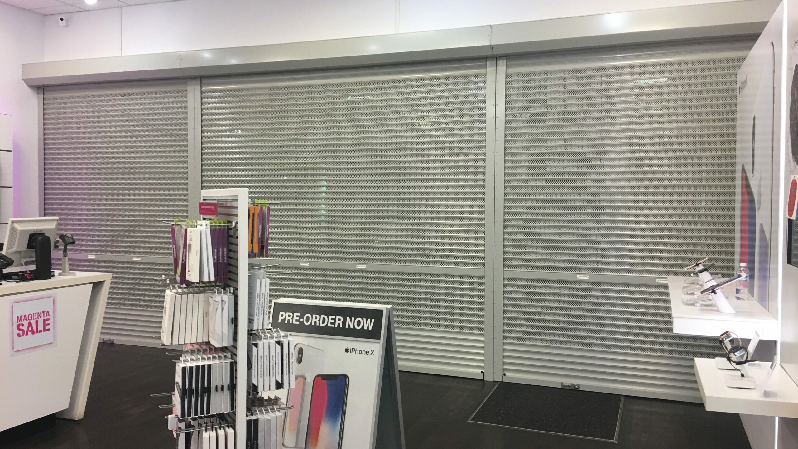 An interior view of a retail store with closed metal shutters at the entrance. To the left, there is a display table with various mobile phone accessories and a promotional sign for pre-orders. The store has a modern design with bright lighting.