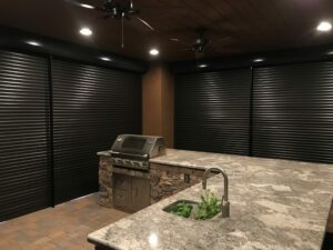 An indoor outdoor kitchen featuring a granite countertop, a built-in grill with a stone facade, and large black rolling shutters. The space is illuminated with ceiling fans and overhead lights, creating a modern appearance.