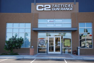 Exterior view of the C2 Tactical Gun Range building, featuring a large sign with the name, glass entrance doors, and displays for firearms and an indoor range. Surrounding landscaping includes trees and shrubs.