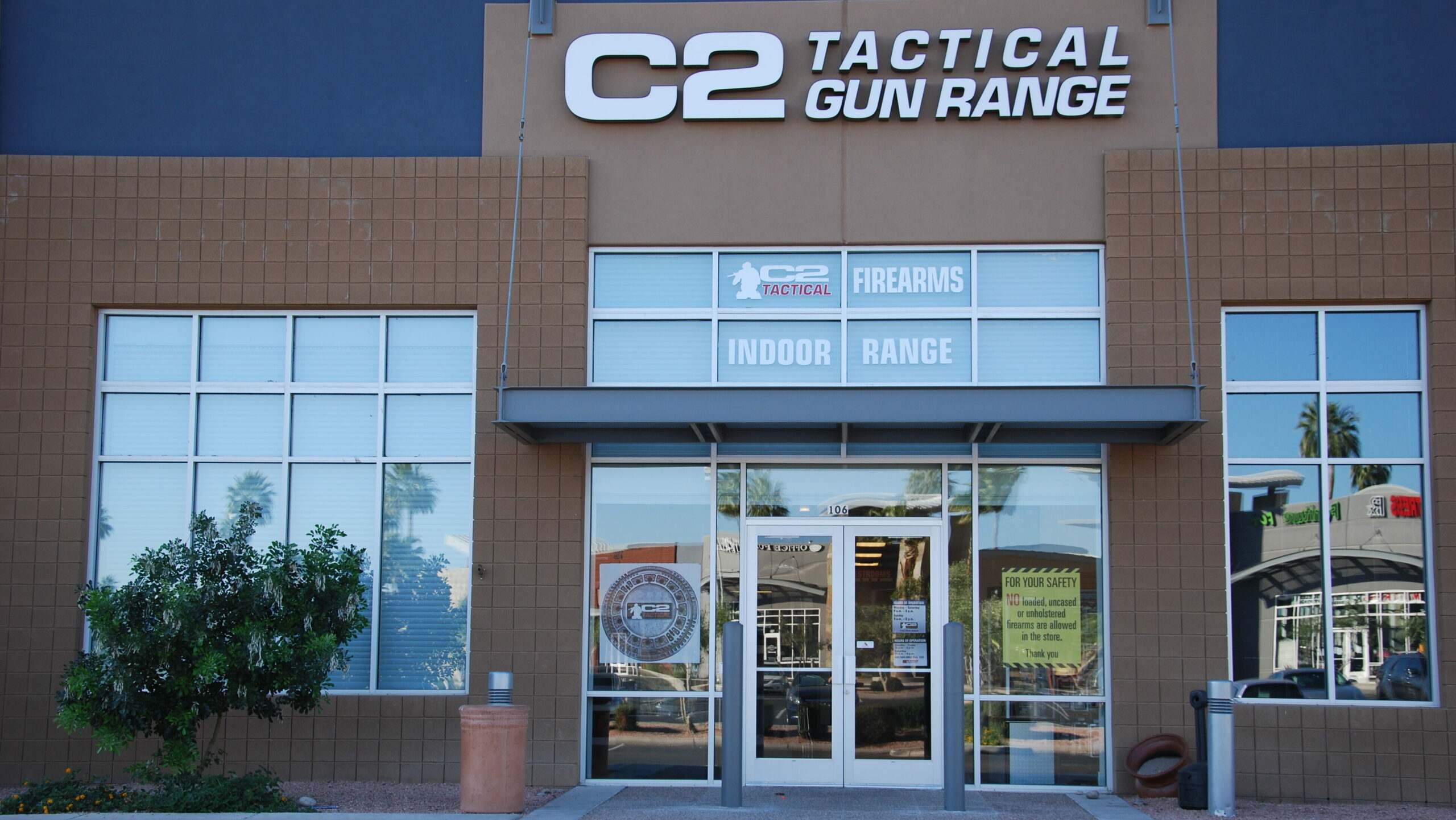 Exterior view of the C2 Tactical Gun Range building, featuring a large sign with the name, glass entrance doors, and displays for firearms and an indoor range. Surrounding landscaping includes trees and shrubs.