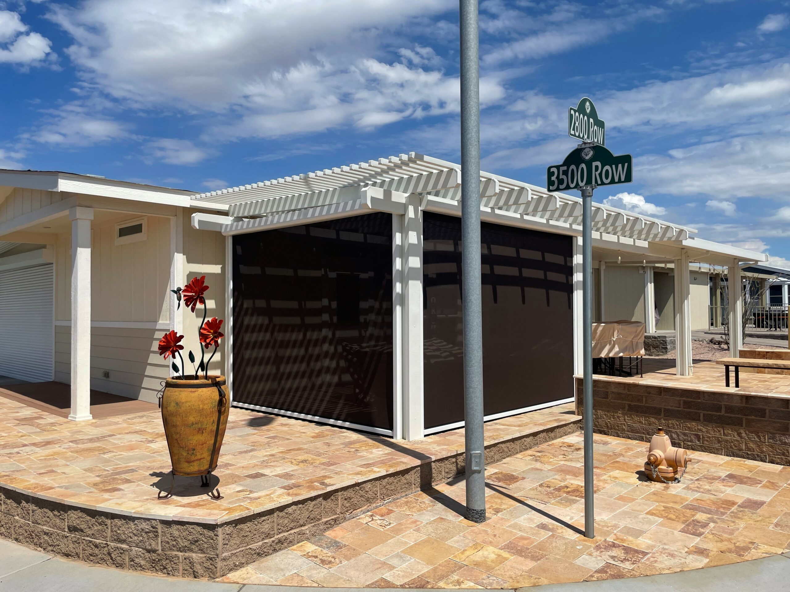 A residential building with a pergola and a dark screen enclosure, featuring a decorative pot with red flowers in front. A street sign indicates u00223500 Rowu0022 nearby, under a blue sky with scattered clouds.