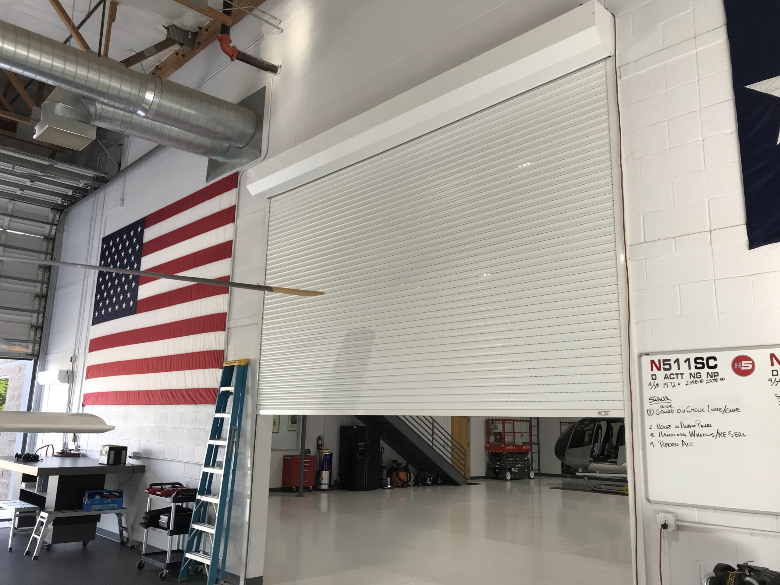 A large white rolling door is closed in an aircraft hangar. An American flag hangs on the wall next to the door. A ladder stands nearby, and various items are visible in the spacious hangar.