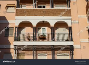 A view of a beige apartment building with a balcony. The balcony features ornate black railings and a planter with flowers. Light streams through the windows, emphasizing the architectural details.