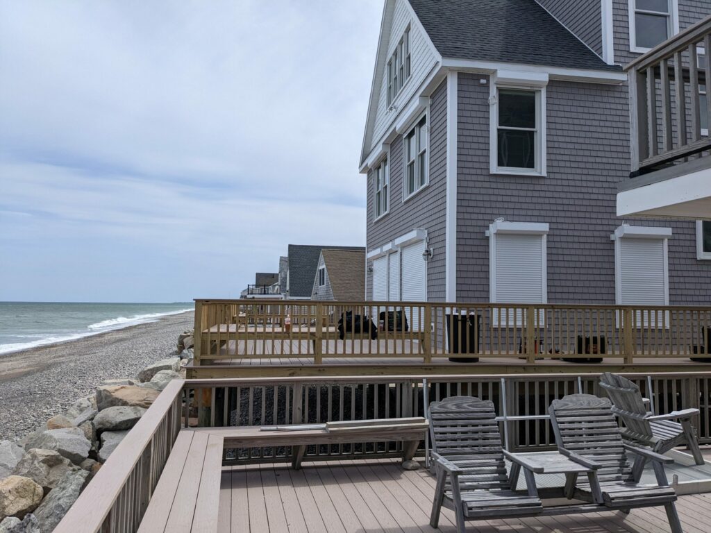Coastal view from a deck featuring wooden chairs, a beach, and gray houses along the shoreline. The ocean is visible with gentle waves, and light clouds are in the sky.