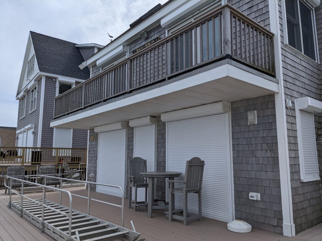A gray shingle-style beach house with a second-floor balcony. Below, there are three closed roll-up window shutters and a small outdoor table with chairs. Wooden decking is visible in the foreground.