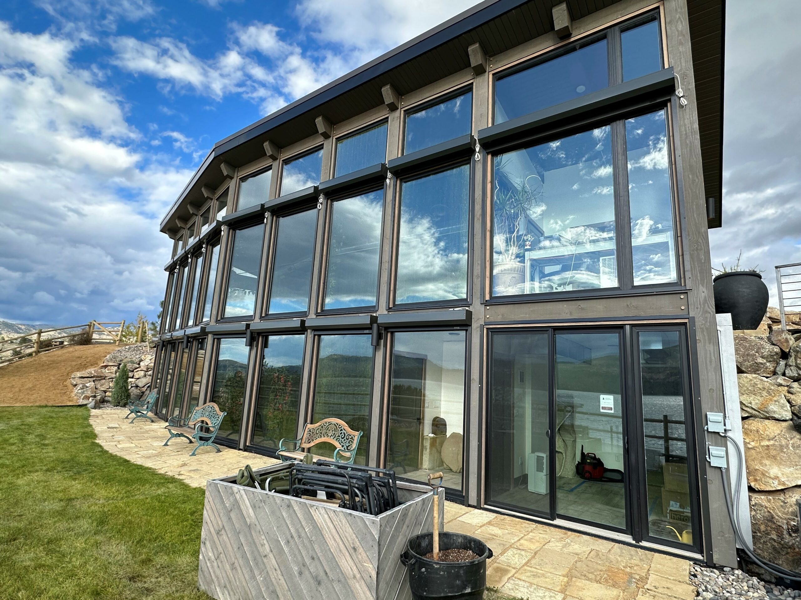 A modern building with large glass windows, set against a cloudy sky. The exterior features a combination of stone and wood, with a stone pathway leading to the entrance. Outdoors, there are chairs and a container with various items.