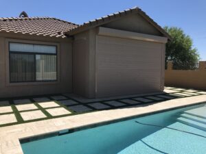 A swimming pool adjacent to a beige house with a tiled roof. The house features a large window and a roll-up door. Green grass patches separate the stone pathway leading from the pool to the house.