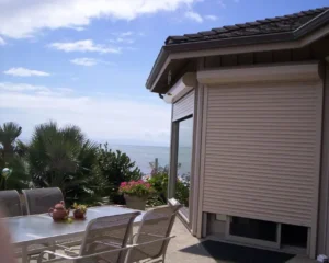 A patio area with a dining table and chairs, surrounded by plants, overlooks a view of the ocean under a partly cloudy sky. The side of a house with a rolled-down shade is visible.