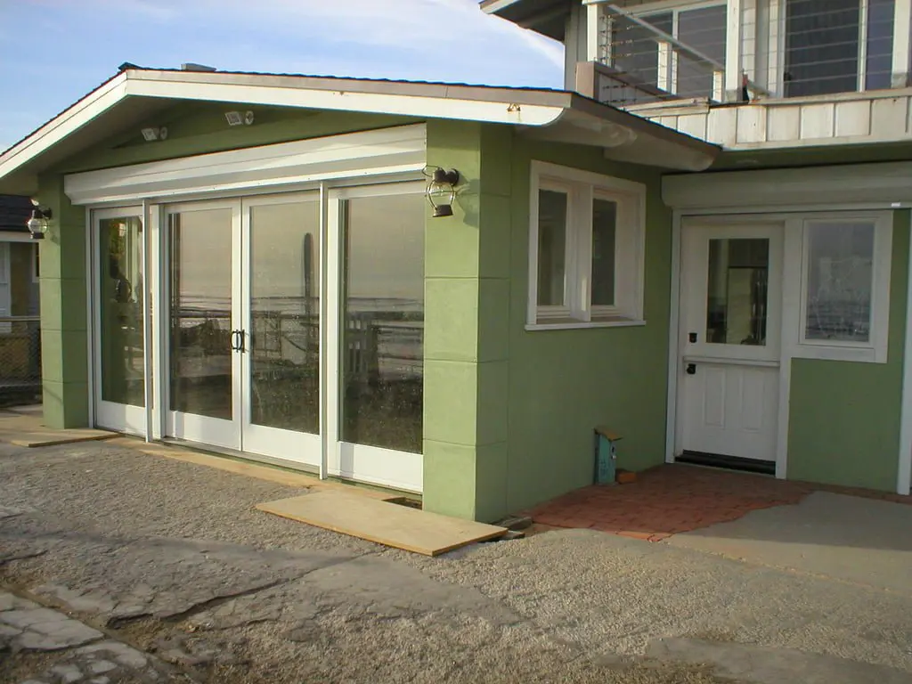 A green house with large glass sliding doors, featuring a small entrance door on the right. The surrounding area is paved with a concrete path, and the roof is slanted.