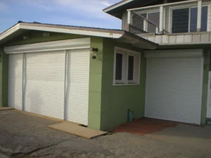 Two garages with white roll-up doors attached to a green house. One garage door is slightly smaller than the other, and the house has a second floor with multiple windows.