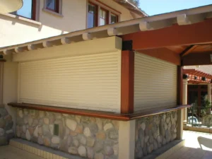 A closed roll-up shutter on a stone wall, part of a modern outdoor area with a wooden overhang. Nearby, there are windows and patio furniture visible in the background.