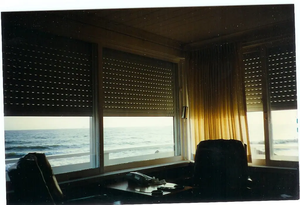 An interior view of a room with two large windows showing the ocean, partially covered by Venetian blinds. A wooden desk with a telephone is in the foreground, and sunlight filters through curtains.