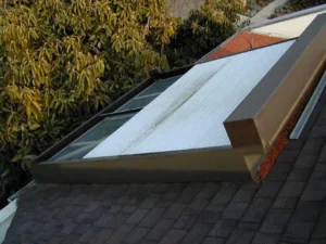 A roof section featuring a sloped skylight and a frame, surrounded by green foliage. The skylight has a white cover, and the roofing material has a dark shingle texture.